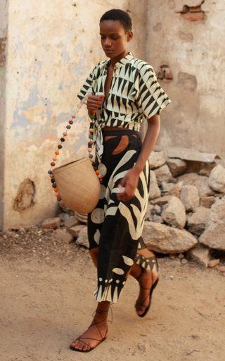 a stylish woman is walking outdoors in an africaninspired outfit. she is wearing a black and white geometric print shirt and skirt, and brown leather sandals. she carries a beige woven bag with colorful beaded straps. the setting is rustic, with stone walls and ground.