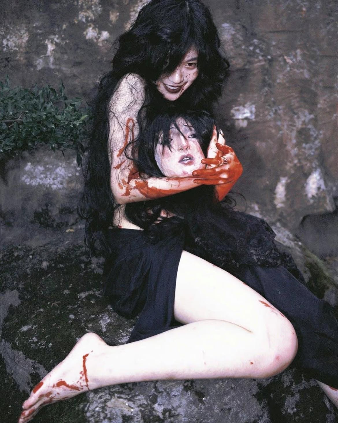 a morbid and disturbing image shows a woman with long, dark hair holding the bloody, severed head of another woman. the woman holding the head has a sinister smile and pale skin. blood is splattered on her hands, arms, and the head she's holding. she is sitting on a rock. the scene is set outdoors, against a backdrop of rocks and sparse vegetation. the overall tone is dark, eerie, and violent, characteristic of the horror genre.