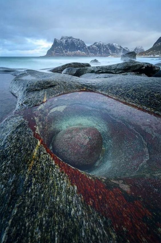 a tide pool filled with crystal clear water sits nestled amongst the rugged rocks of a beach. the water is calm and still, reflecting the mountains in the distance. the rocks surrounding the pool are a mix of dark grey and red, creating a beautiful contrast to the blue water. the scene is peaceful and serene, and it evokes a sense of awe at the beauty of nature.