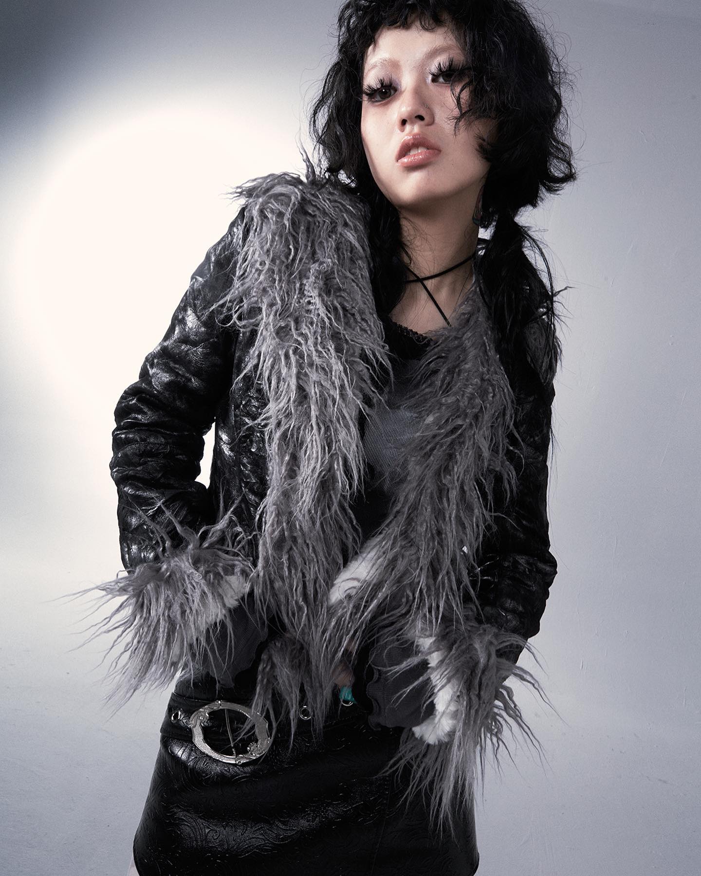 a young woman with dark, short, curly hair is modeling a stylish black leather jacket adorned with a thick gray faux fur trim.  she's also wearing a black leather mini skirt and a simple silver belt with a circular buckle. her makeup is dark and accentuates her eyes.  the overall style is edgy, blending elements of punk and goth fashion. the background is a simple, muted gray, focusing attention on the model and her outfit.  the image is a professional studio shot, likely for a fashion magazine or website.