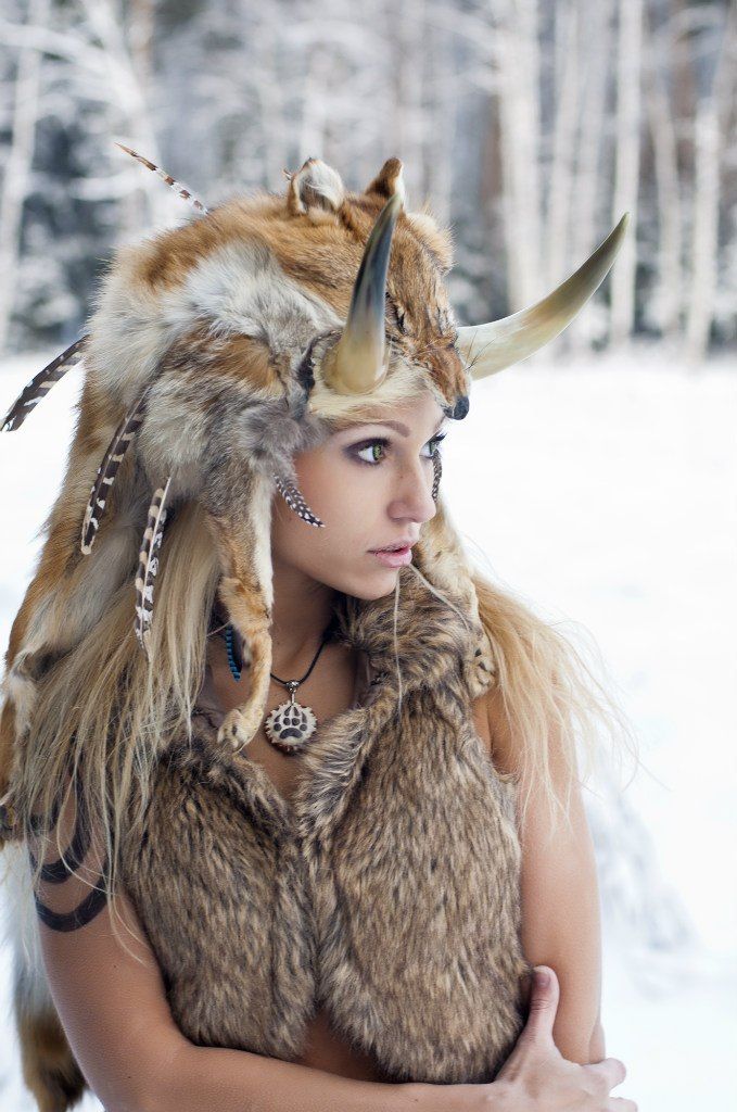 a beautiful blonde woman with a fur hat and a fur vest, standing in a wintery forest. she is looking away from the camera with a soft expression on her face. the woman is wearing a fur hat with feathers and horns attached to it, and her vest is made of animal fur. the background is a snowy forest, with the trees covered in snow. the woman is dressed in a fashion style reminiscent of native american traditions.