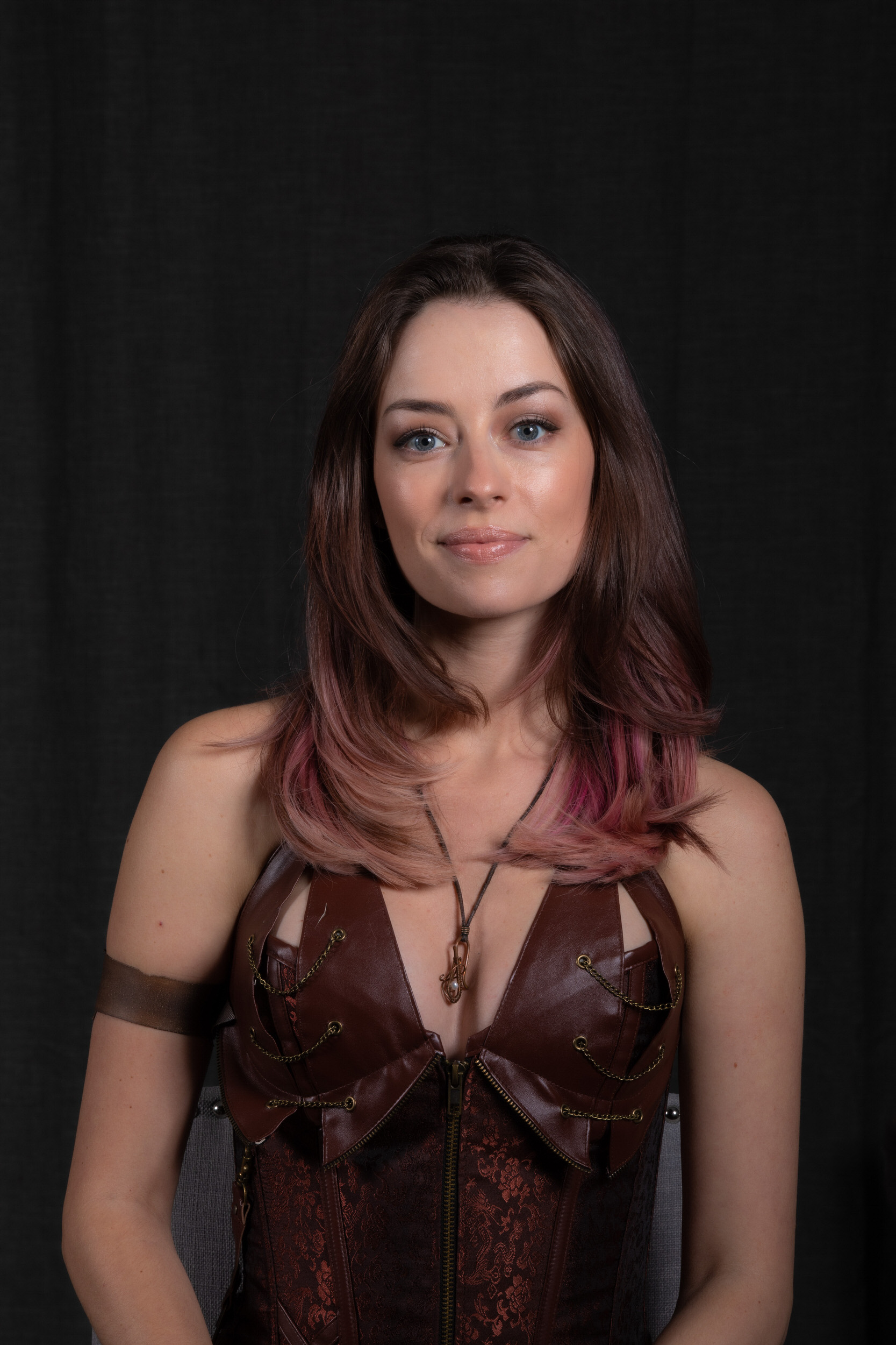 a woman with pinktinted brown hair is sitting in front of a black backdrop. she is wearing a brown steampunkstyle corset top and a necklace with a pendant. she is looking at the camera with a slight smile on her face.