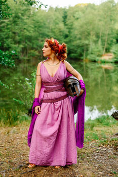 a fulllength outdoor shot of a redhead woman in a long, flowing purple dress.  she is wearing a style reminiscent of ancient greek or roman clothing, including a belted chiton or stola. she carries a darkcolored, what appears to be a corinthian helmet, under her arm.  she is standing by a lake in a forest setting, her gaze directed away from the camera.  the overall impression is of a historical reenactment or cosplay.