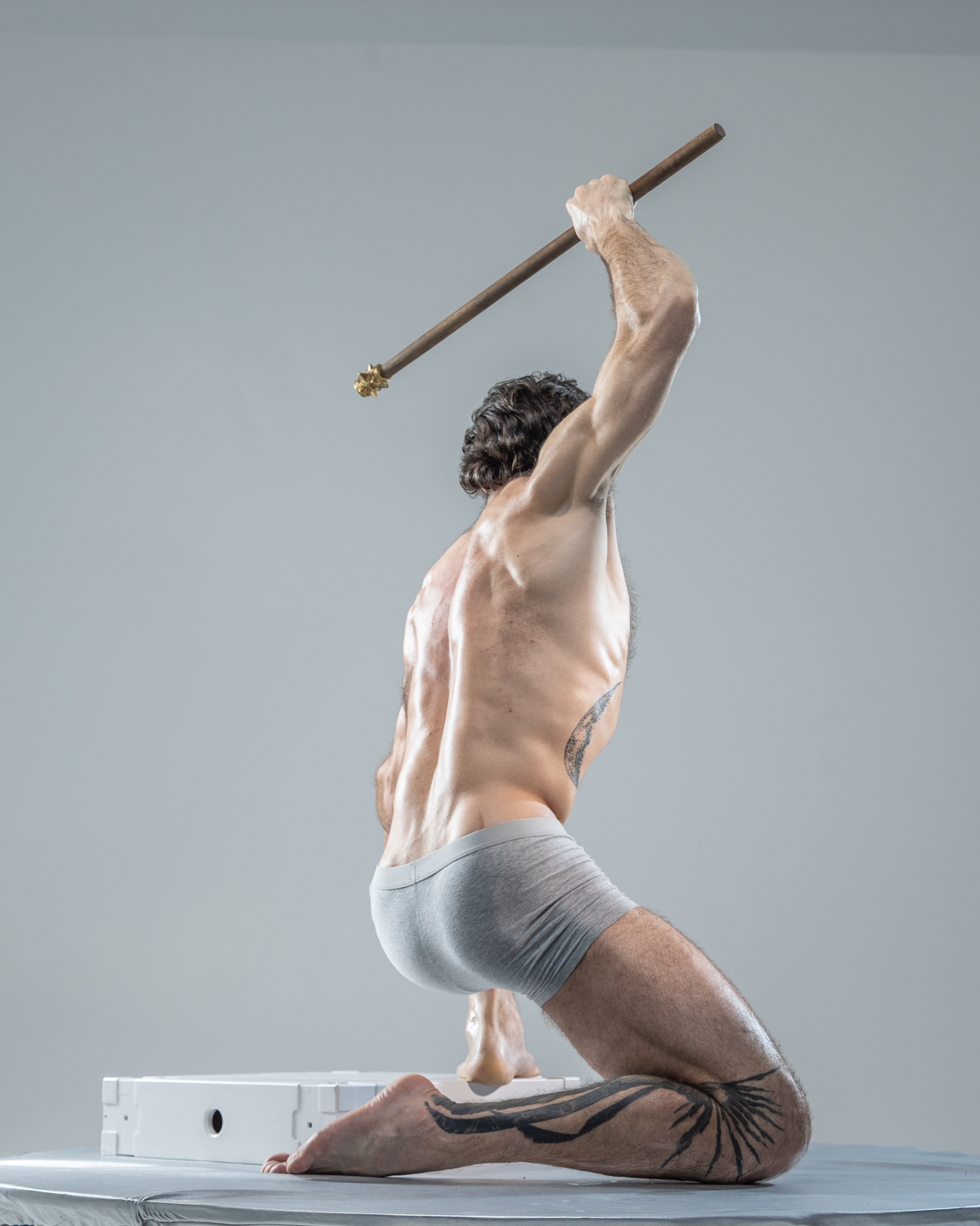 a muscular man with tattoos is posed against a light grey background in a dynamic stance. he is holding a staff above his head, seemingly about to strike. he is wearing grey underwear and his body is illuminated by studio lighting, highlighting his musculature. the image is a back view taken from a low angle, emphasizing his powerful posture. the overall composition evokes a sense of strength, power, and athleticism, drawing inspiration from classical art and warrior archetypes.