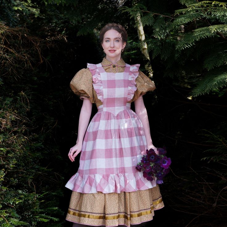 a woman in a vintageinspired dress stands in a forest. she is wearing a pink and white gingham dress with a white ruffled apron. she is holding a bouquet of purple anemones. the background is a lush green forest. the lighting is soft and natural.