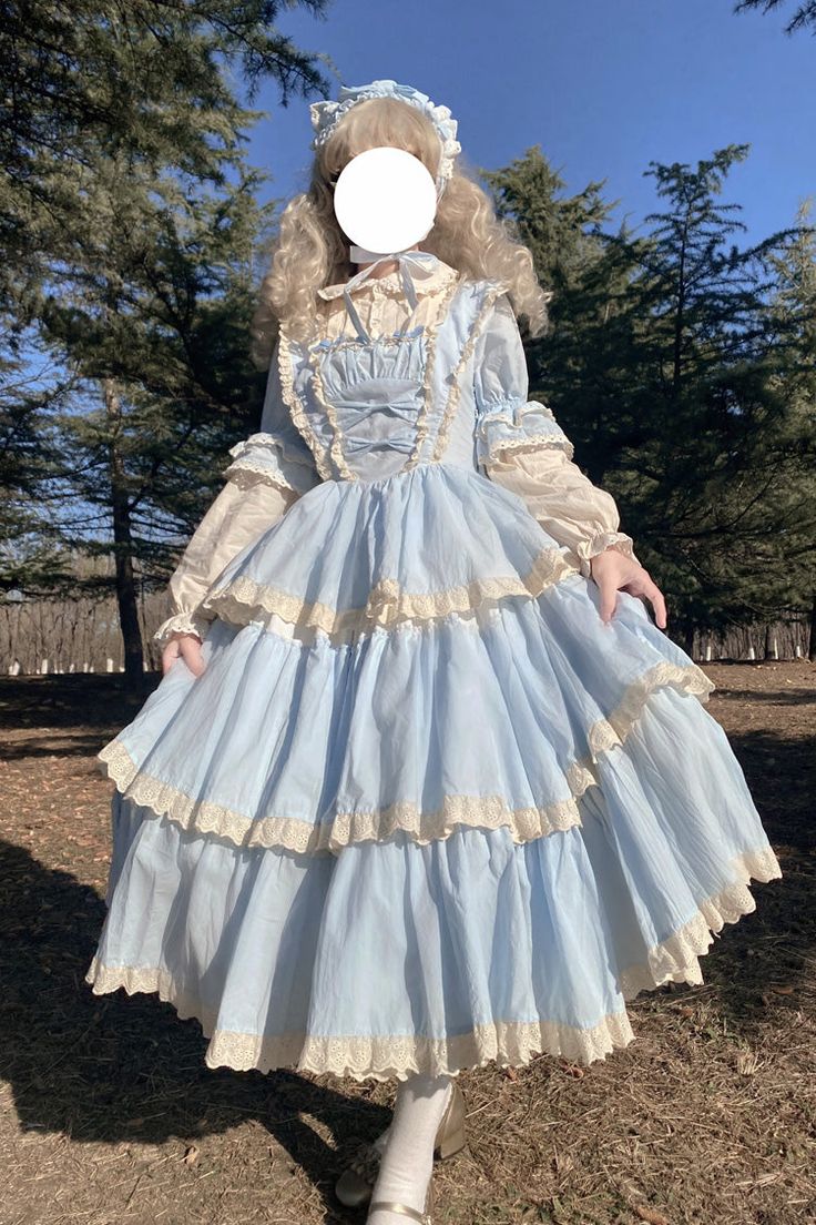 a woman wearing a blue and white lolita dress and bonnet stands in a park. the dress is tiered and has lace details. the bonnet is white with lace. the woman is standing on grass and there are trees in the background. the photo is taken from eye level and the lighting is natural and sunny.