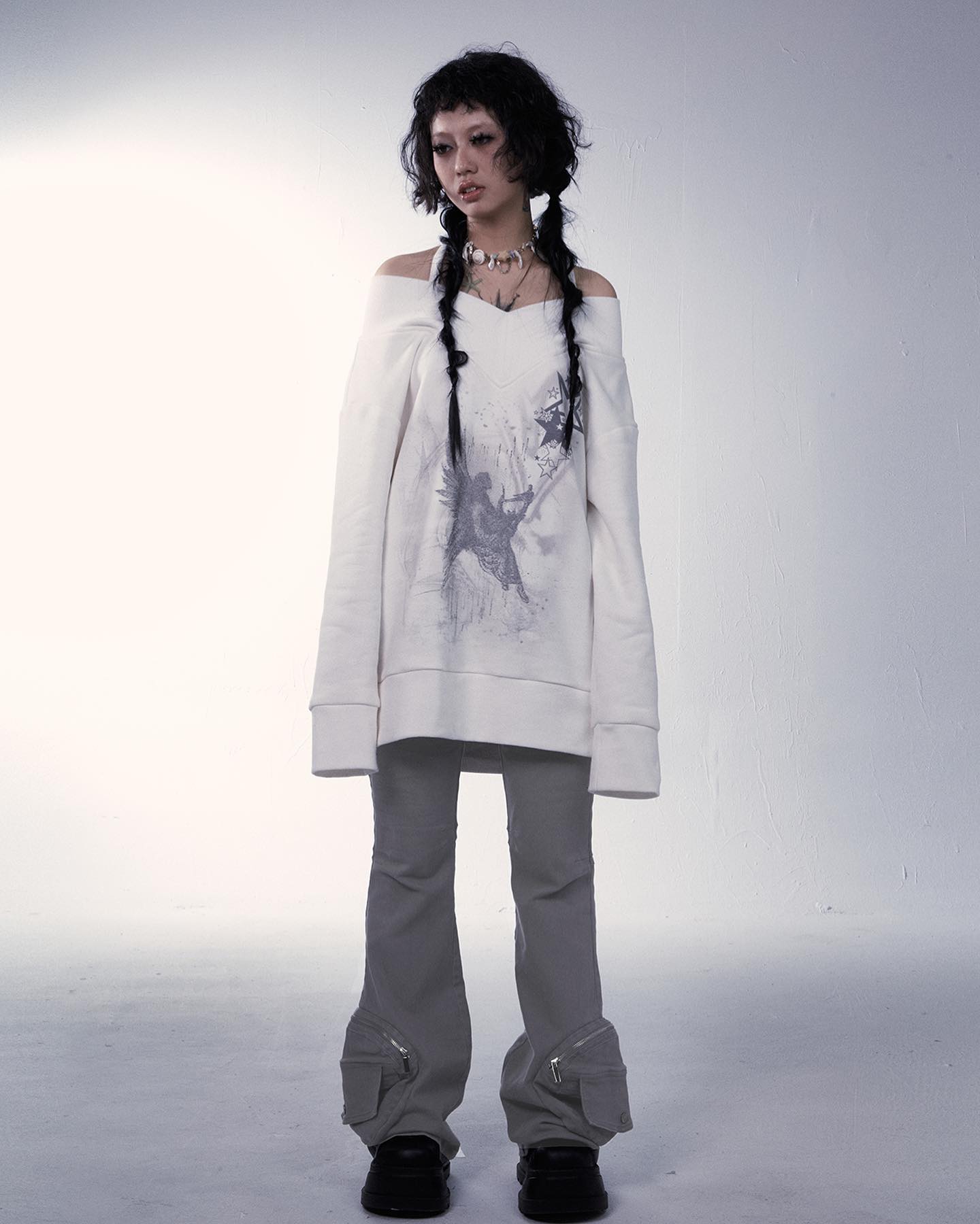 the image showcases a young woman, possibly a model, dressed in an offtheshoulder, oversized creamcolored sweatshirt featuring a grey graphic print that seems to depict an angel or similar figure.  she's wearing light grey cargo pants with zip details at the ankles. her dark hair is styled in two braids. she's accessorized with a simple necklace. the background is a plain, lightgrey studio setting, emphasizing the clothing. the overall aesthetic is a blend of streetwear and alternative/gothic styles.  the muted color palette and minimalist backdrop create a clean and visually appealing image.