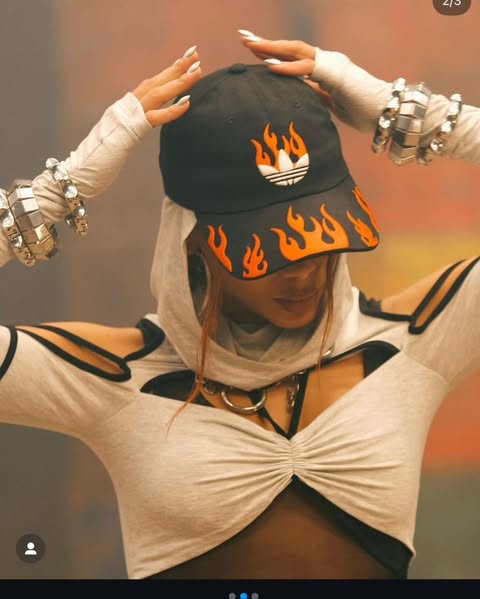 a woman is posing indoors wearing a black adidas hat with flame designs, a white hoodiestyle top with black accents, and silver jewelry, including bracelets and rings. she is looking down.
