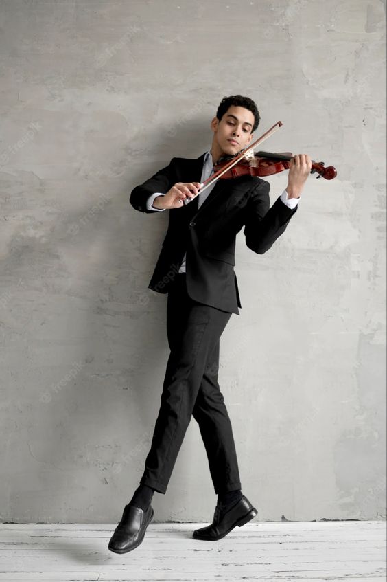a young man dressed in a black suit stands against a gray concrete wall, playing a violin. he is elegantly posed, with one leg extended and his eyes closed, fully absorbed in the music. the studio setting and his formal attire suggest a professional or artistic context.