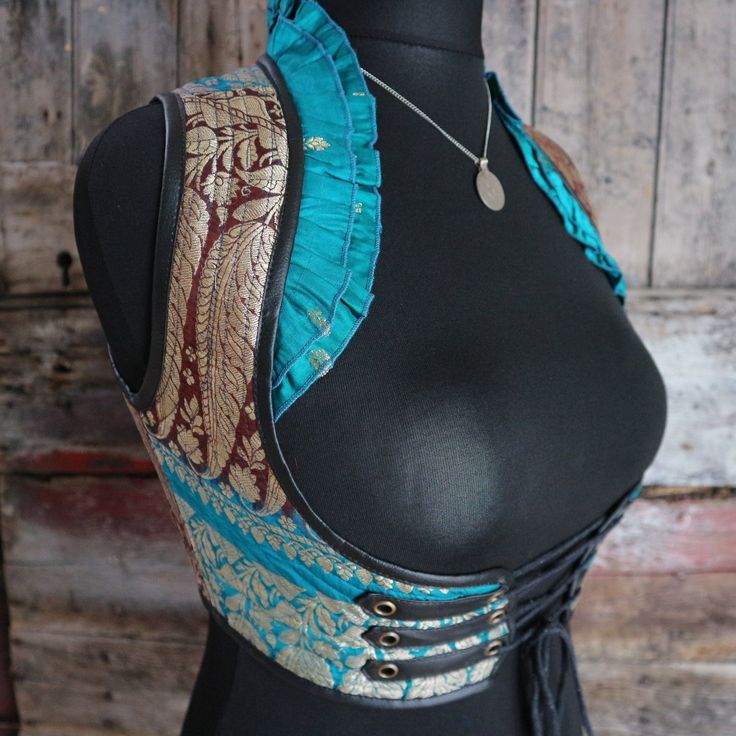 a photograph of a mannequin wearing a bustierstyle garment. the garment is made of turquoise and gold brocade fabric with leather accents and ruffles. it is a vestlike piece that has a lacedup corset style on the sides. the mannequin also has a necklace with a silver pendant. the mannequin is set against a rustic wooden background.