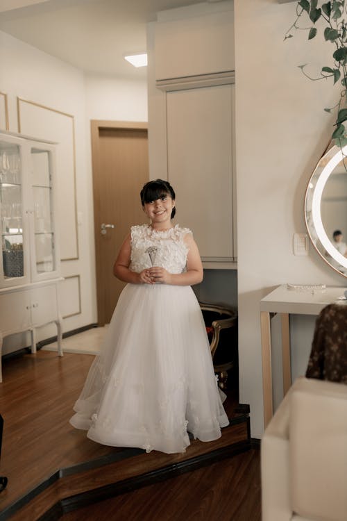 a young girl in a beautiful white flower girl dress stands in a nicely decorated room. she holds a small bouquet in her hands and smiles at the camera. the room features elegant furniture, including a white cabinet and a vanity with a mirror. the floor is made of hardwood. the girl's dress is long, white and tulle, with floral details. the atmosphere is bright and airy, typical of a wedding or special event setting.