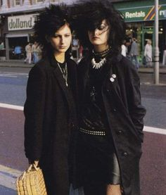 two young women in gothic style clothing stand together on a city street in the 1980's. one woman is wearing a black coat with a black leather skirt and the other woman is wearing a black coat with a black leather skirt. they both have black hair and are posed looking directly at the camera.  they are both wearing necklaces and both have a rock and roll or punk style about their appearance.