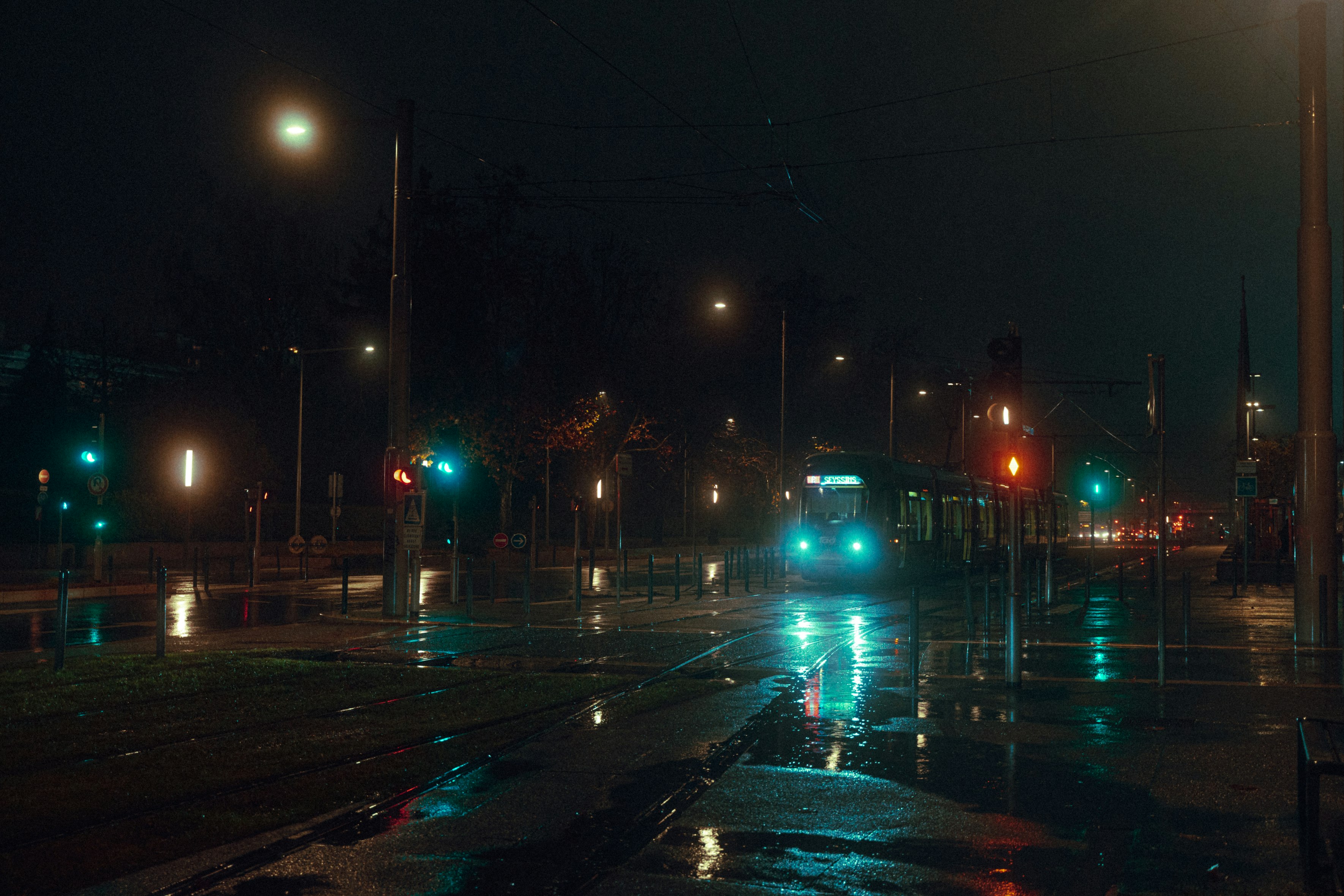 a night shot of a streetcar traveling down a wet street in the city. the street is illuminated by streetlights and the tram's headlights. the reflection of the lights can be seen in the puddles on the road. the scene is moody and atmospheric.