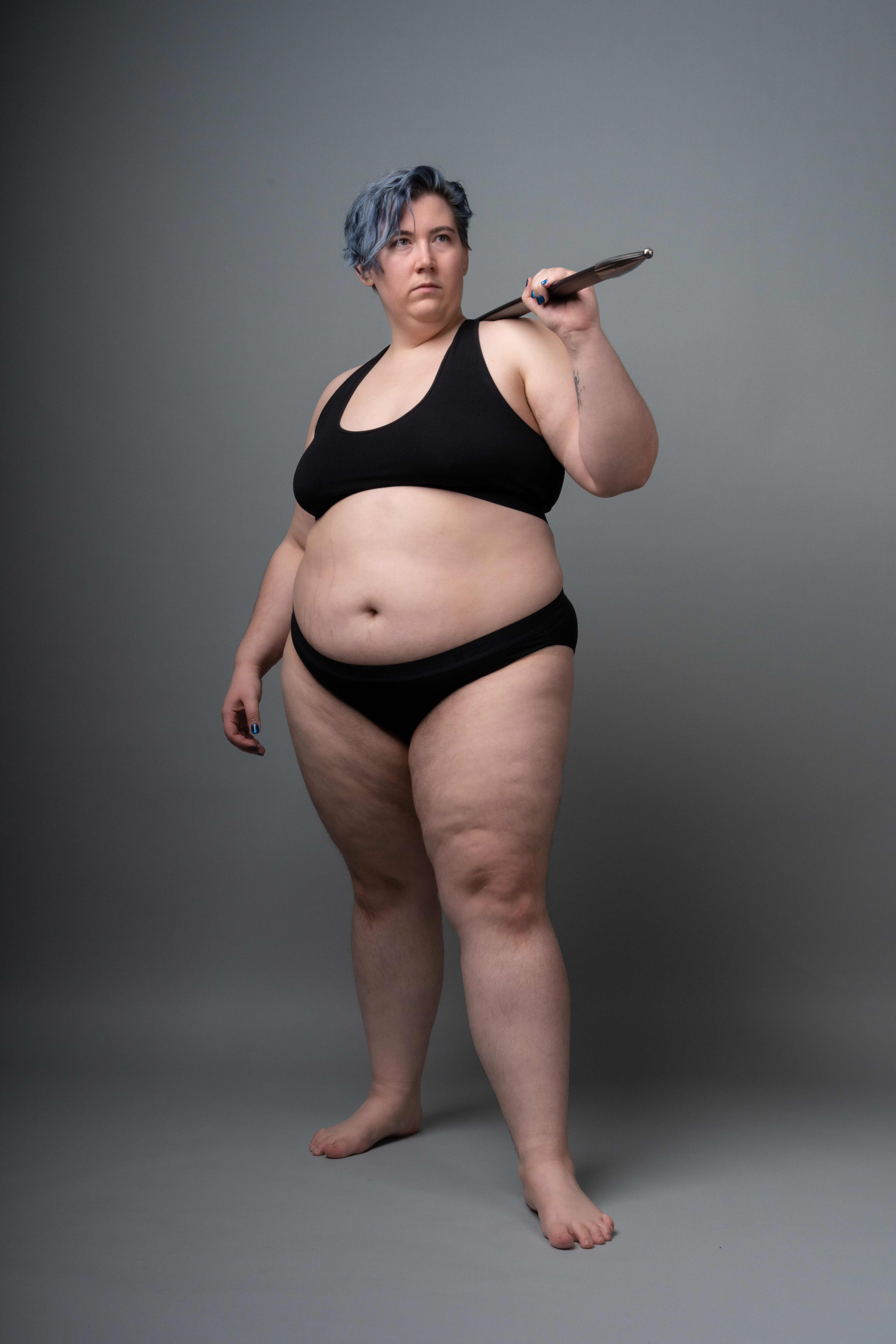 a full studio shot of a plussize woman with blue hair wearing a black bra and underwear. she is barefoot and holding a weapon over her shoulder. the woman is looking away from the camera. the background is gray.