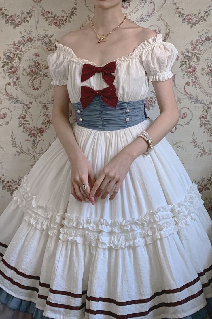 a woman is shown from the chest down, wearing an elegant white lolita fashion dress. the dress has an off the shoulder neckline with puffy short sleeves, a blue basque waist with a bow at the center and red ribbon accents, and a long, flowy skirt with layers of ruffles and red ribbon details. the background of the image is a floral wallpaper.
