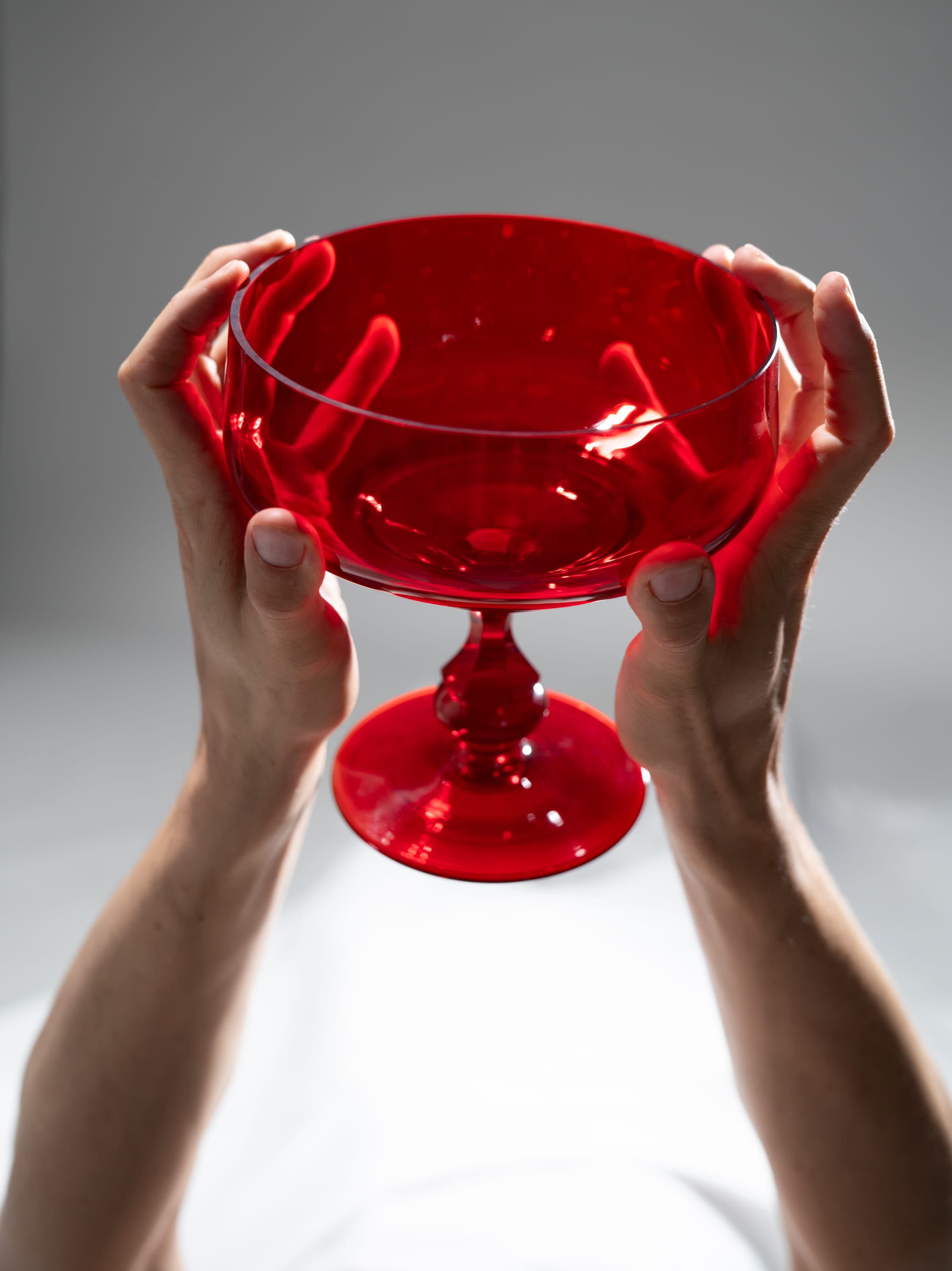 a pair of hands delicately holds a vibrant red glass goblet against a minimalist gray background. the goblet's translucent nature allows light to pass through, illuminating the hands and creating a captivating interplay of color and shadow.