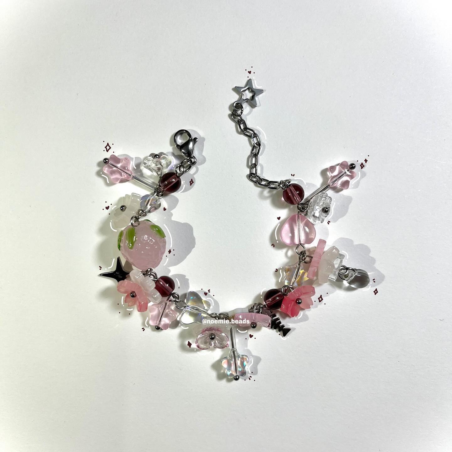 a beaded bracelet with a chain is displayed on a white surface. the bracelet features a variety of beads in shades of pink and clear, along with flowershaped charms and starshaped accents. the chain is silver in color and has a star charm at the end.