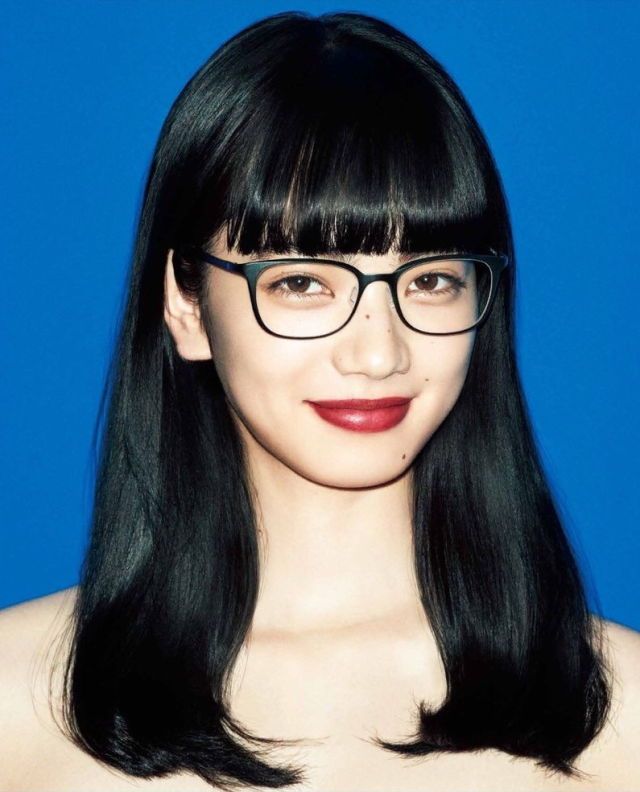 a portrait of a young asian woman with black hair and bangs, wearing glasses, looking at the camera. she is wearing red lipstick. the background is a blue wall.