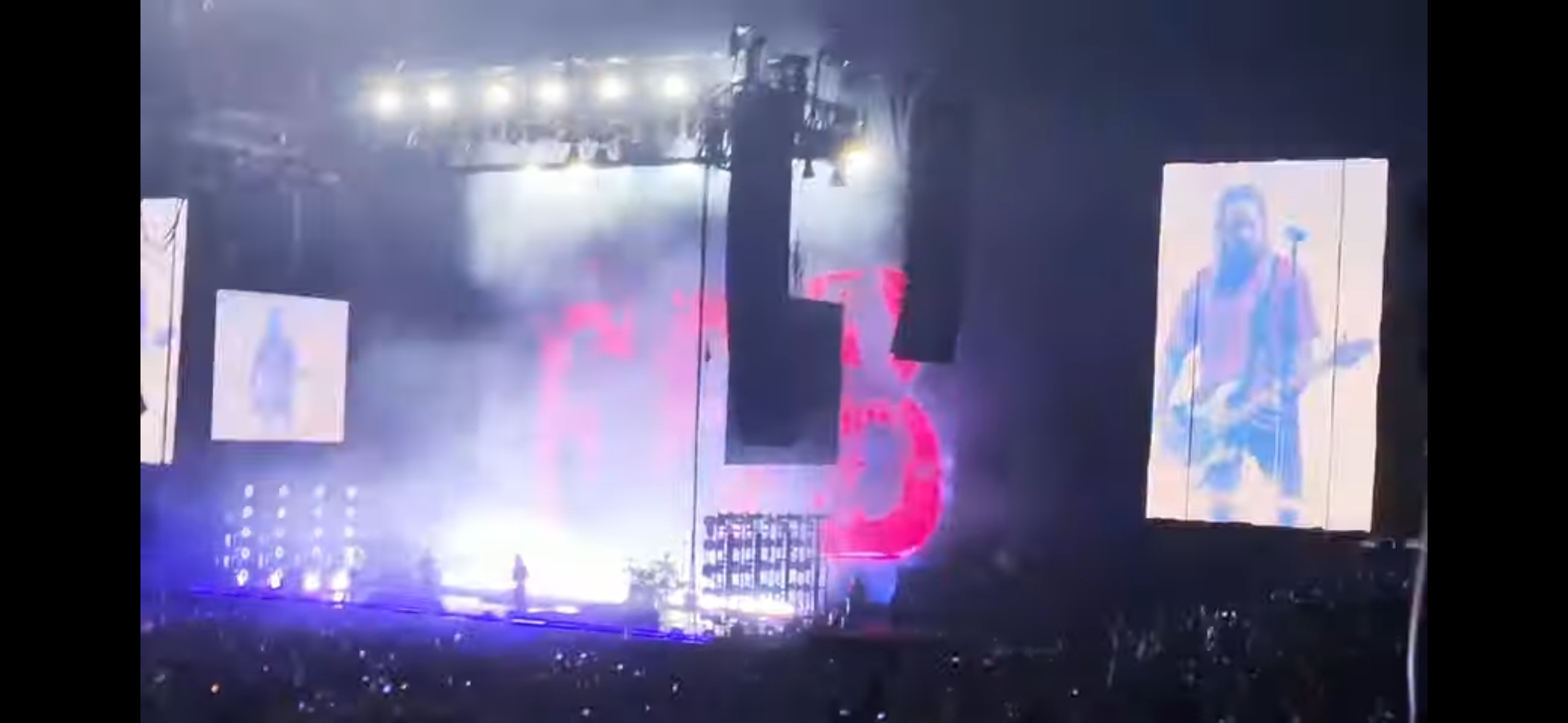 a concert stage with a large screen displaying a guitar player. the stage is lit up with lights and there is a large crowd in the audience. the screen behind the guitar player has two giant glowing red letters on it.