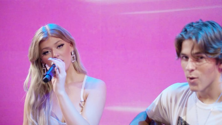a woman with long blonde hair is singing into a microphone while wearing earrings and a white top. a man with brown hair is to her right wearing a white tshirt. they are both in front of a pink background.