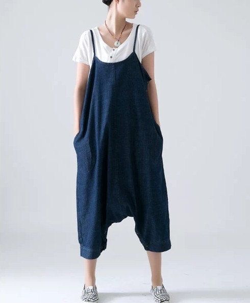 a woman is standing indoors, wearing a white tshirt underneath a denim jumpsuit with spaghetti straps and pockets. she is also wearing a necklace and loafers. the jumpsuit is a loose, casual fit.