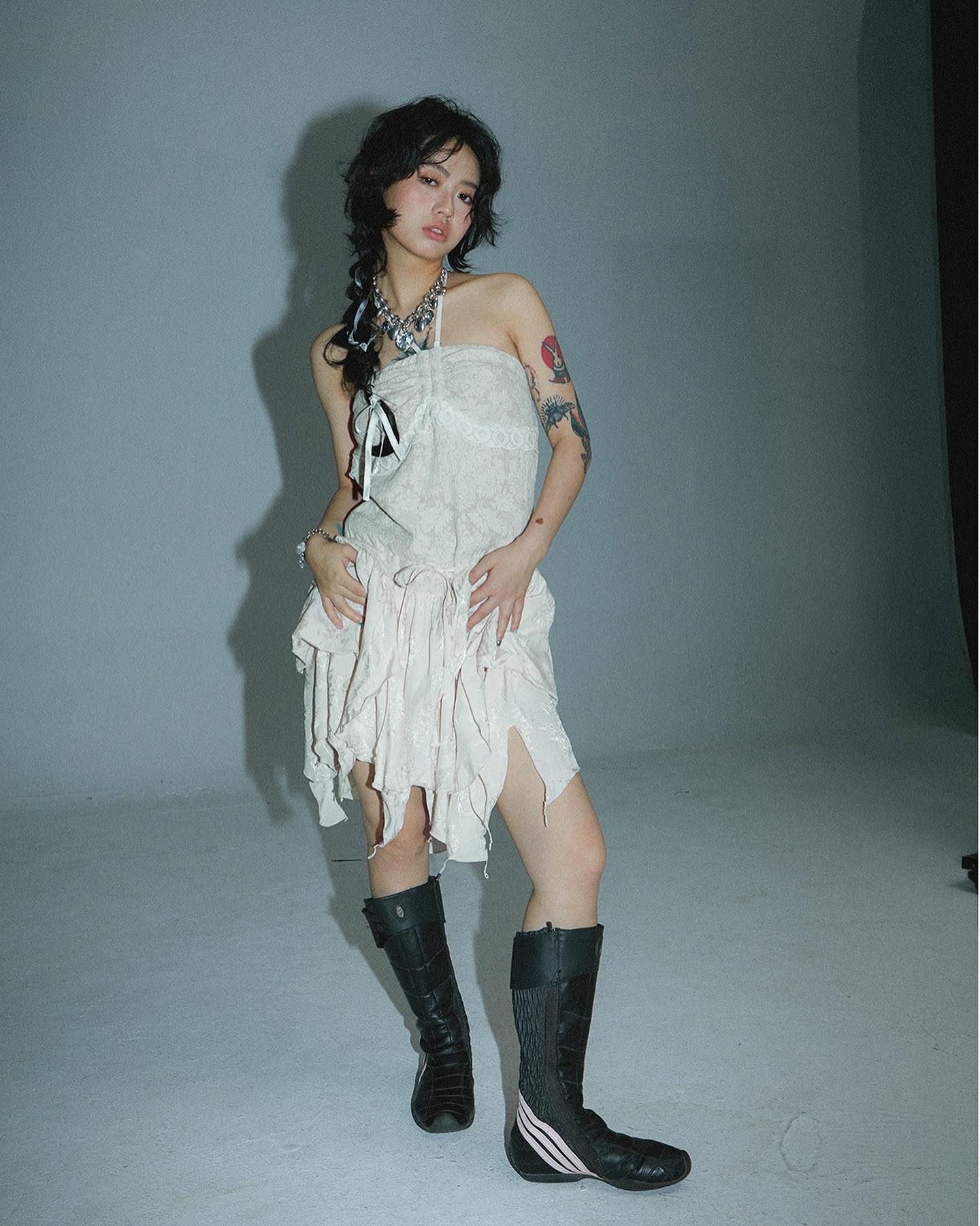 a young woman with dark hair styled in a layered cut poses in a studio. she's wearing a light beige, ruffled, sleeveless mini dress, black high boots with a stripe detail, and a silver necklace. she has visible tattoos on her arm and shoulder and is looking directly at the camera with a slightly serious yet stylish expression. the background is a plain light gray. the overall style is edgy and fashionable, with a hint of grunge aesthetic. the image has a slightly grainy, filmlike quality.