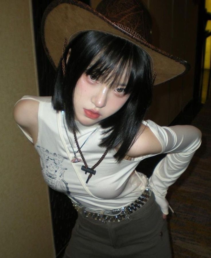 a young woman is taking a selfie in a hallway mirror. she is wearing a white shirt, brown pants, a brown cowboy hat, and a rhinestone belt. she has short black hair.