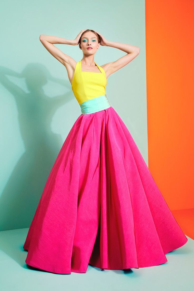 a woman is posing in a studio wearing a vibrant outfit consisting of a yellow halter top, a turquoise belt, and a large, full pink skirt. she has her hands behind her head. the background is a split of turquoise and orange.