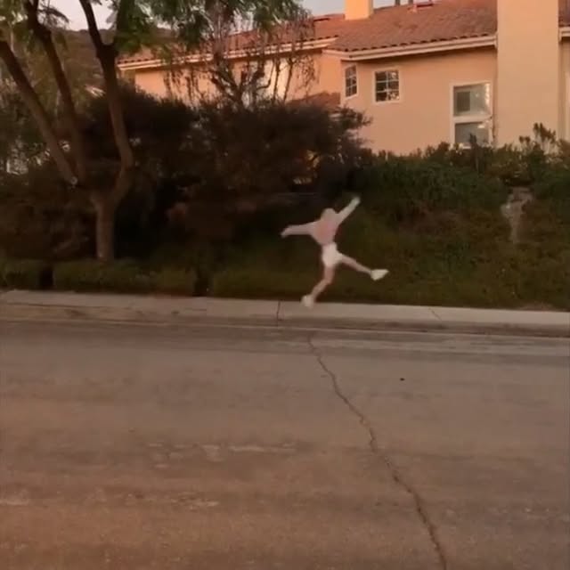 a person in a light pink longsleeved shirt and white shorts jumps on a sidewalk in front of a residential area during the daytime. a twostory building is visible in the background behind bushes and a tree.