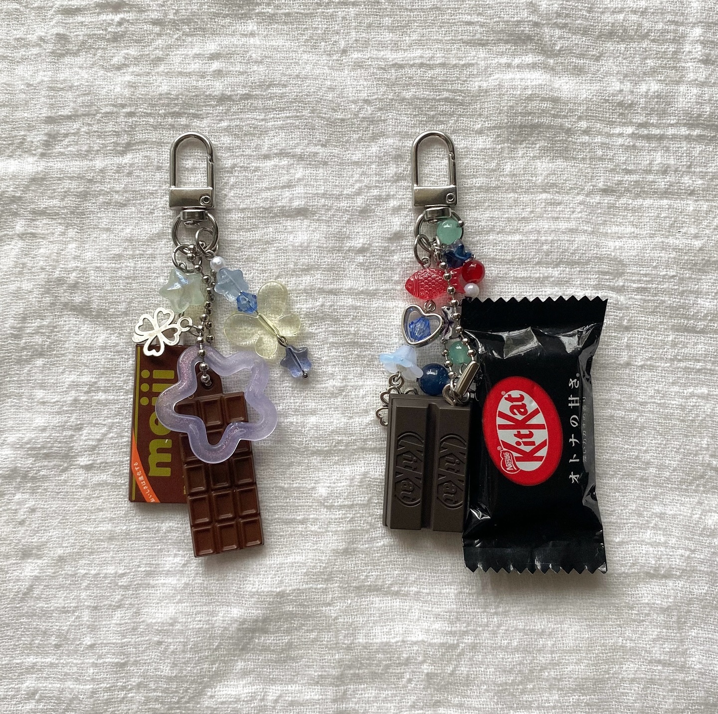 two keychains with chocolatethemed charms are arranged side by side on a textured white fabric. the keychain on the left features a brown muji chocolate bar charm, a translucent purple star, and a chocolate bar charm. it also has silver charms of a clover and colorful beaded butterfly accents. the keychain on the right features a blackwrapped japanese kit kat charm, a kit kat bar charm, beads and heart shaped charms. both keychains are attached to a silvertoned clasp.