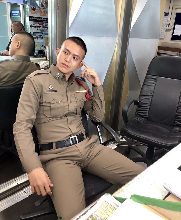 a young asian man in a police uniform sits in a chair in an office setting, looking serious and thoughtful. he is wearing a brown uniform with a badge, black belt, and brown pants. he is sitting with one hand on the desk and the other on his chin, and he is looking off to the side as if he is contemplating something. the office is a modern one with a desk, a chair, and some documents on the desk.