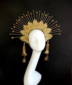 a goldcolored headdress is displayed on a white mannequin head against a black background. the headdress features a sunburst design with metallic spokes radiating outward from a central floral motif. two gold tassels hang down from the sides of the headdress, adding to its ornate and eyecatching appearance. the mannequin head is positioned facing forward, showcasing the full beauty and intricate details of the headdress.