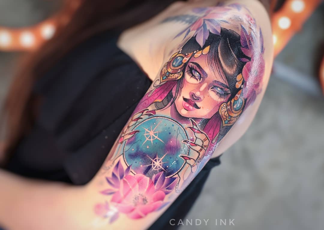 a closeup shot of a colorful tattoo on someone's upper arm. the tattoo depicts a woman with purple hair and white eyes, adorned with gold and jewels, holding a crystal ball that shows a galaxy within. she is surrounded by pink flowers.  the tattoo is done in a neotraditional style with bold lines and smooth shading. the artist's watermark, "candy ink," is visible in the bottom right corner.