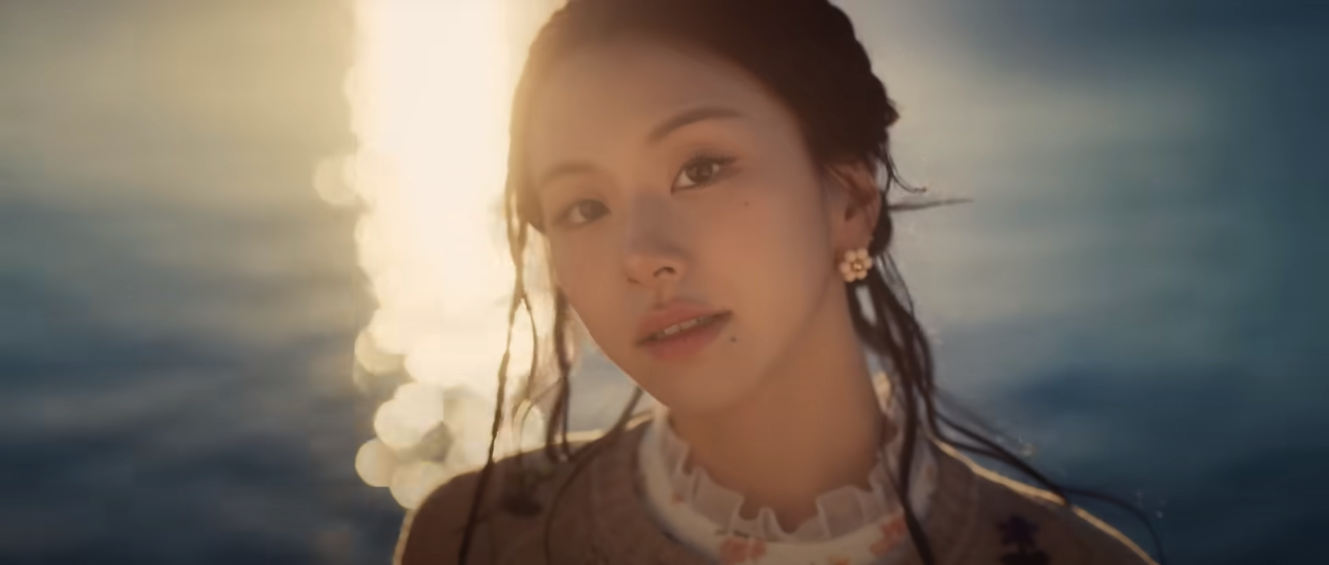 the image is a closeup portrait of a young asian woman with her hair pulled back standing on a beach at sunset. the golden light of the setting sun illuminates her face and creates a warm, ethereal glow. the background is slightly blurred, emphasizing the woman's serene expression and the peaceful atmosphere. she is wearing a creamcolored sweater with floral embroidery and pearl earrings. the overall mood is one of beauty, tranquility, and connection with nature.