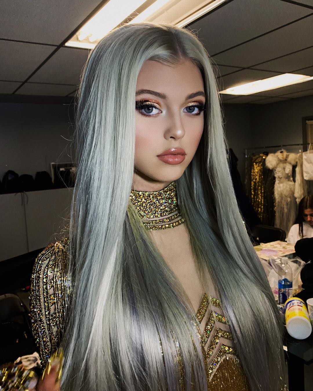 a woman with long, light green hair, wearing a gold dress and gold jewelry. she has makeup on, including eye makeup and lipstick. she appears to be wearing a wig.