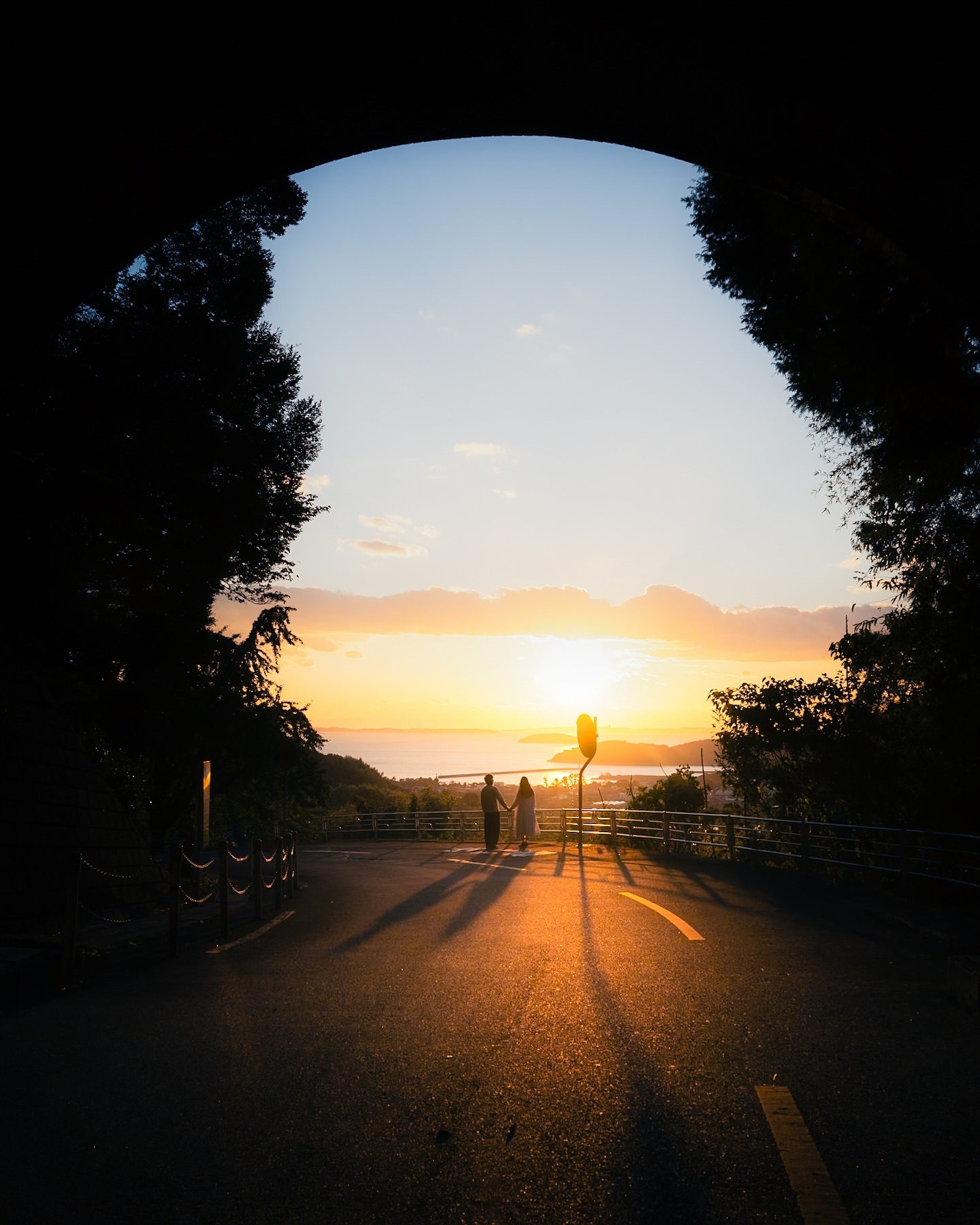 the image shows a silhouetted couple holding hands, standing on a road at the end of a tunnel.  the tunnel's archway frames a breathtaking sunset over a calm ocean.  trees line the road, creating a natural border. the warm light of the setting sun casts long shadows across the road, enhancing the romantic and serene atmosphere. the overall composition creates a dramatic and visually appealing scene.