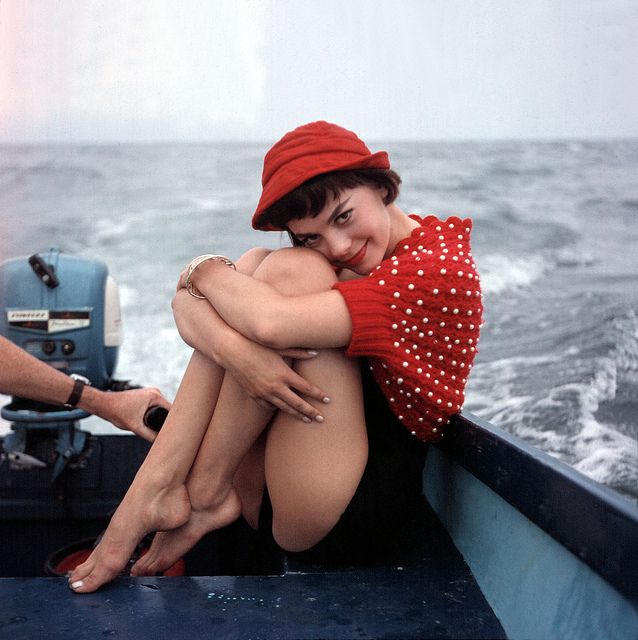 a young woman in a red beaded sweater and black bottoms sits on the bow of a small motorboat.  she is hugging her knees and smiling softly at the camera.  the ocean stretches out behind her, and the sky is a hazy blue.