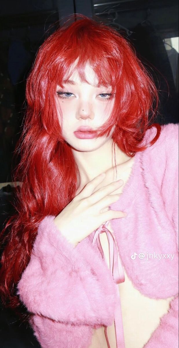 a young woman with vibrant red hair and a stylish fringe poses for a portrait. she wears a soft pink, fuzzy, cropped cardigan. the lighting is soft and diffused, giving her a dreamy look.  the image feels stylish and trendy.