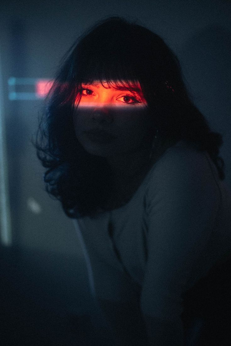 a woman with dark hair and a red neon light reflecting in her eyes stares thoughtfully into the distance. her features are barely visible in the shadows, but her eyes shine with a cool and mysterious light. she is dressed in a simple, dark top.