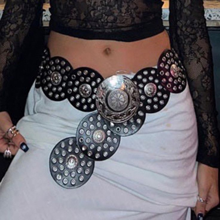 a person wearing a black lace top and white skirt with a wide, decorative belt with large circular metal accents. the belt is positioned around the waist, and the person is wearing multiple rings.