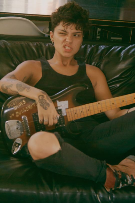 a young woman with short, dark hair sits on a leather couch with a guitar in her lap. she is wearing black tank top and black jeans and playing the guitar. there is a tattoo on her arm and she has a cool, serious expression on her face.