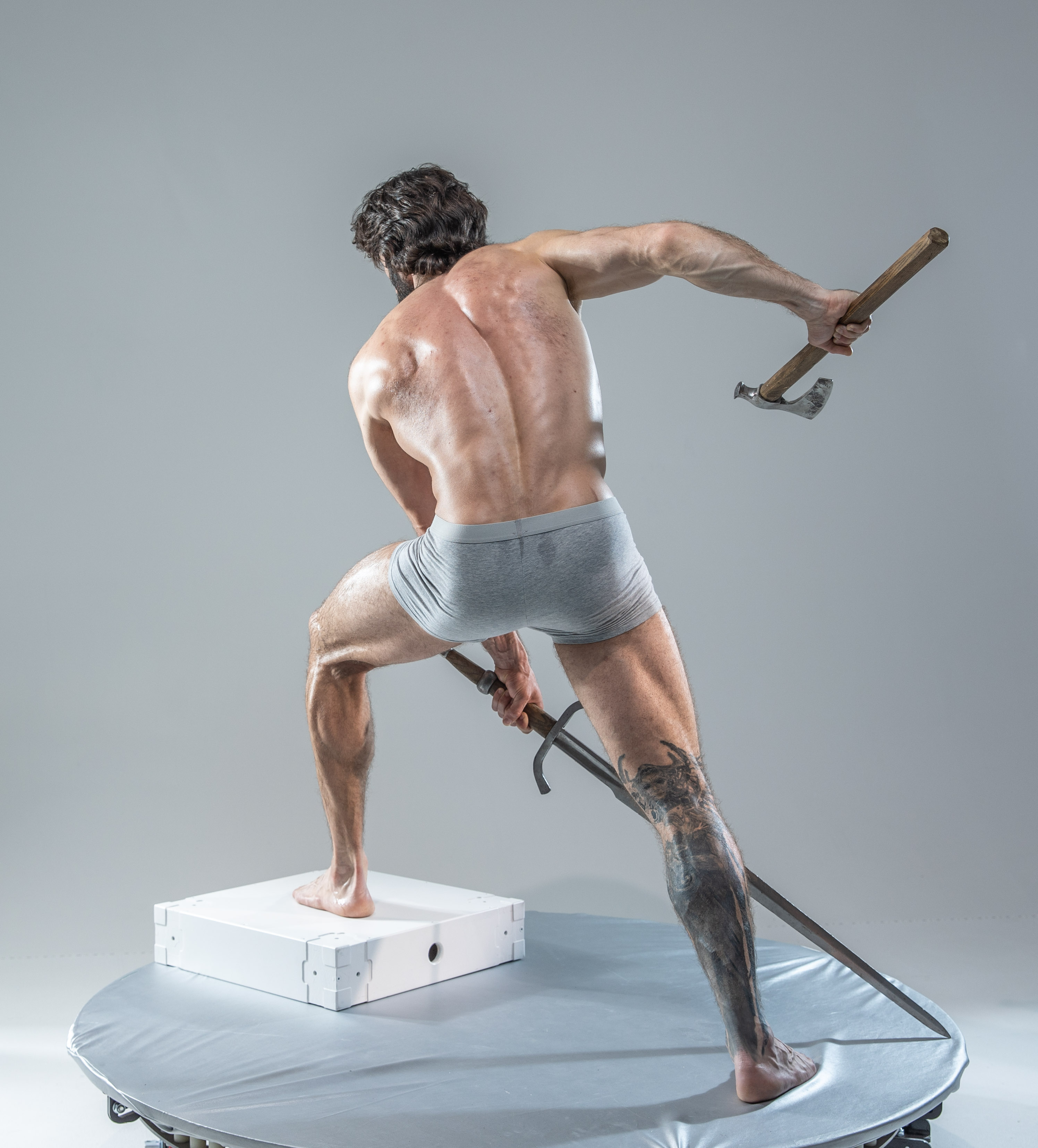a muscular man with dark hair and tattoos is captured midaction in a studio setting against a gray backdrop. he stands on his left leg, right leg extended forward, as if lunging. he holds a sword in his right hand, the blade pointed towards the ground, and an axe in his left hand, raised in the air. he wears only grey underwear, showcasing his physique. the lighting is soft and diffused, highlighting the contours of his muscles. the image evokes a sense of power, strength, and dynamic movement.