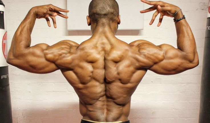 a muscular black man is facing away from the camera, showing off his sculpted back muscles. he is in a gym setting with punching bags visible in the background. the image highlights his impressive physique and the definition of his back muscles.