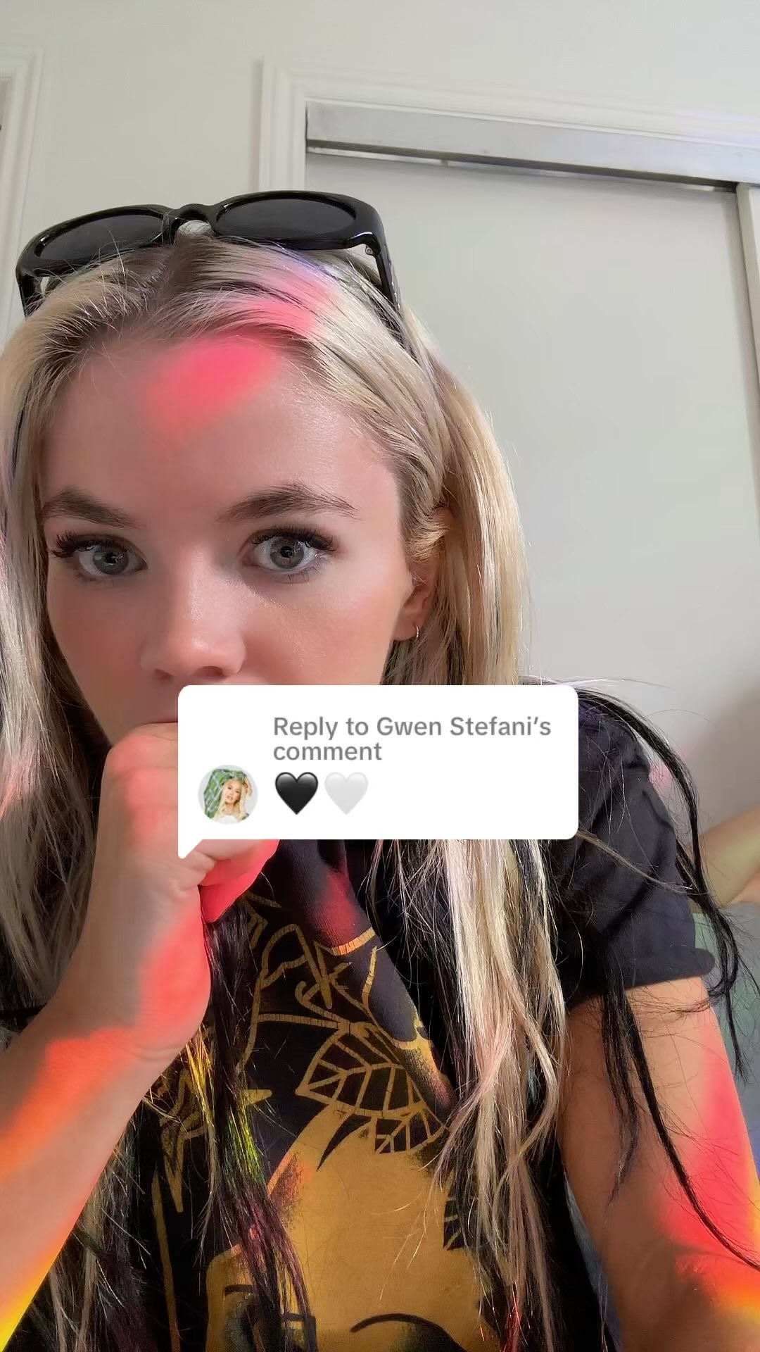 a woman with blonde hair and sunglasses on her head is looking at the camera. there is a comment box that says "reply to gwen stefani's comment" with a picture of a woman with green hair and two hearts next to it. she is wearing a black shirt with a gold design.