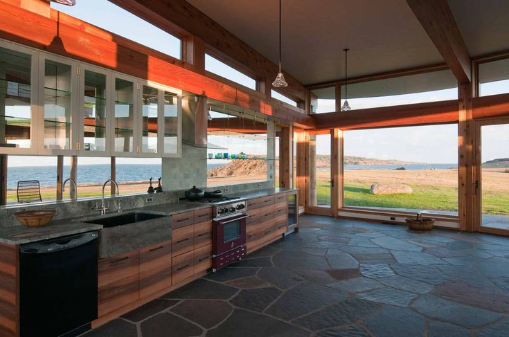 a modern kitchen with wooden beams and cabinets, stone countertops, a sink, dishwasher, and range, with large windows providing an ocean view, stone floor, and pendant lights.
