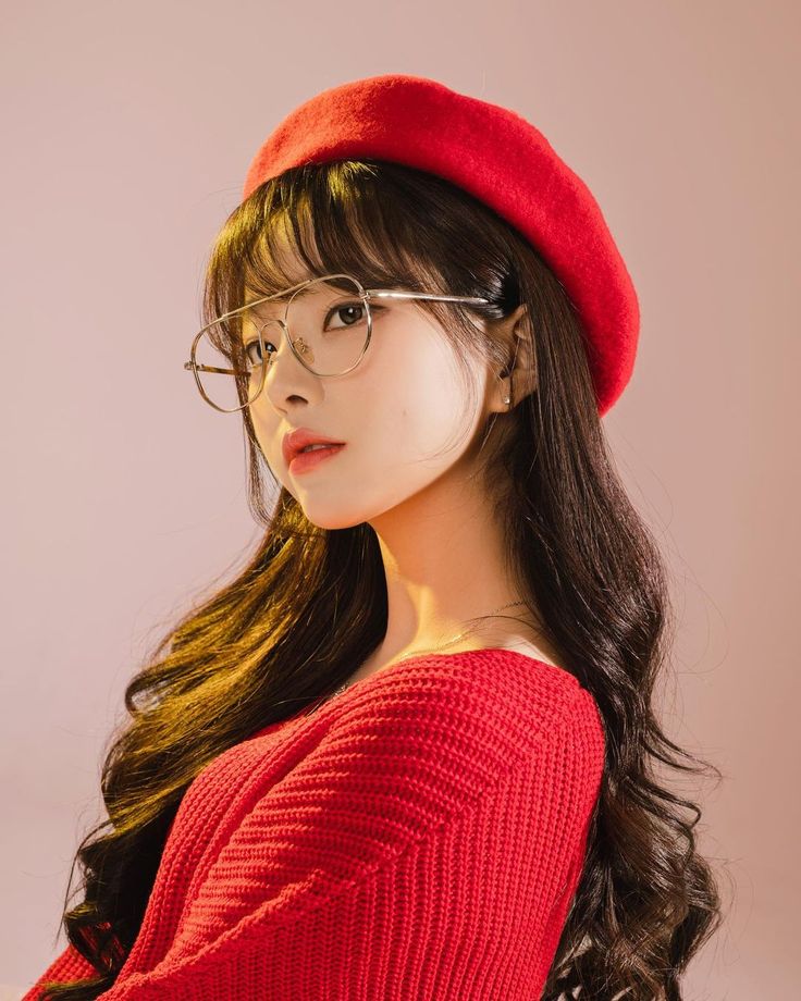 a closeup portrait of a young asian woman with long, wavy dark brown hair. she's wearing a red beret, round silverrimmed glasses, and a red knit sweater. her hair is styled with bangs and her makeup is natural and subtle. the background is a pale pink.  the overall style is elegant and fashionable.