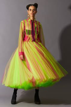 a young woman with short dark hair is wearing a bright and bold fashion outfit. she is standing in a studio setting with a grey backdrop. her dress is a combination of green, yellow, and pink stripes. the skirt is a sheer tulle fabric. the top is a sheer striped buttondown shirt, and the model is wearing a pair of black heeled boots. she is looking forward with a confident expression.