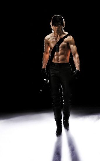 a shirtless man in black leather pants and boots poses confidently with a fierce expression. he is standing on a white platform with dark shadows creating dramatic lines behind him. the background is dark, highlighting his muscular physique and leather attire.