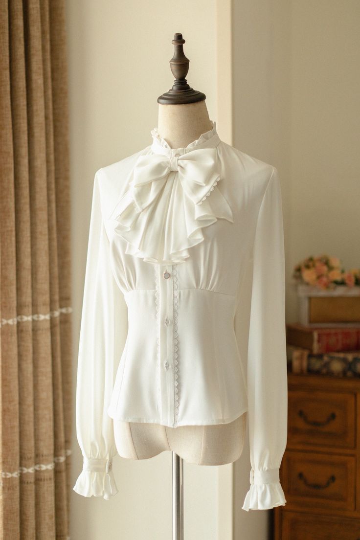 a white, ruffled, victorianstyle blouse is displayed on a mannequin. the blouse has a high neck with a large bow, long sleeves with ruffles at the cuffs, and a fitted bodice with buttons down the front. the background is a creamcolored wall with a white curtain to the left and a white dresser with a floral arrangement on top to the right.
