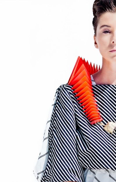 a woman is wearing a black and white striped dress and a red necklace. she is standing against a white background and her eyes are looking at the camera.